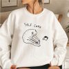 Funny white sweatshirt with a stretching frog cartoon and the text “Self Care” above it.