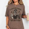 Brown t-shirt with “Cleats & Chaos” skeleton mom graphic holding coffee mug and cleats, with lightning bolt accents.